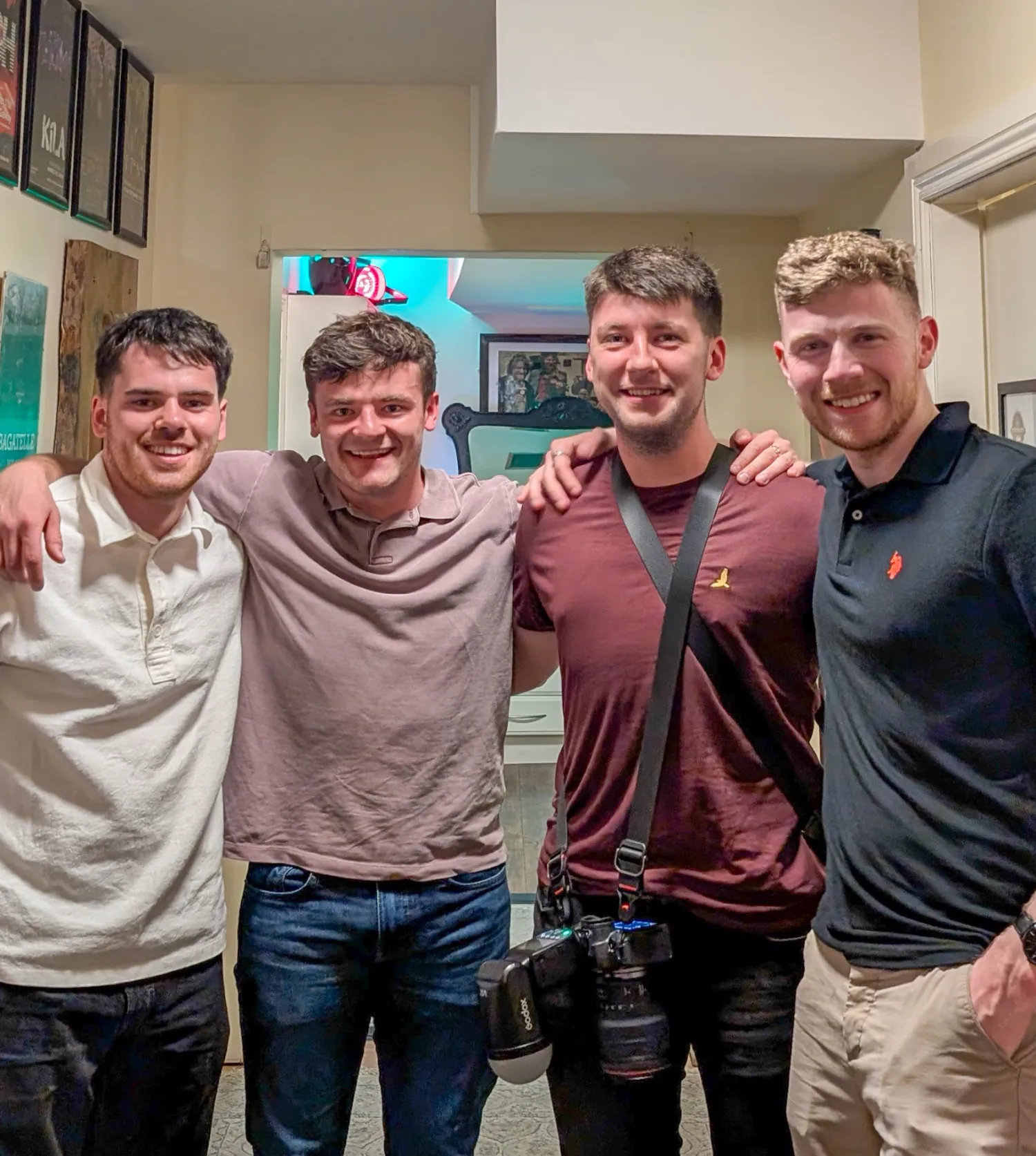 A group of friends stand close together in casual attire, smiling and embracing in what appears to be an indoor setting.