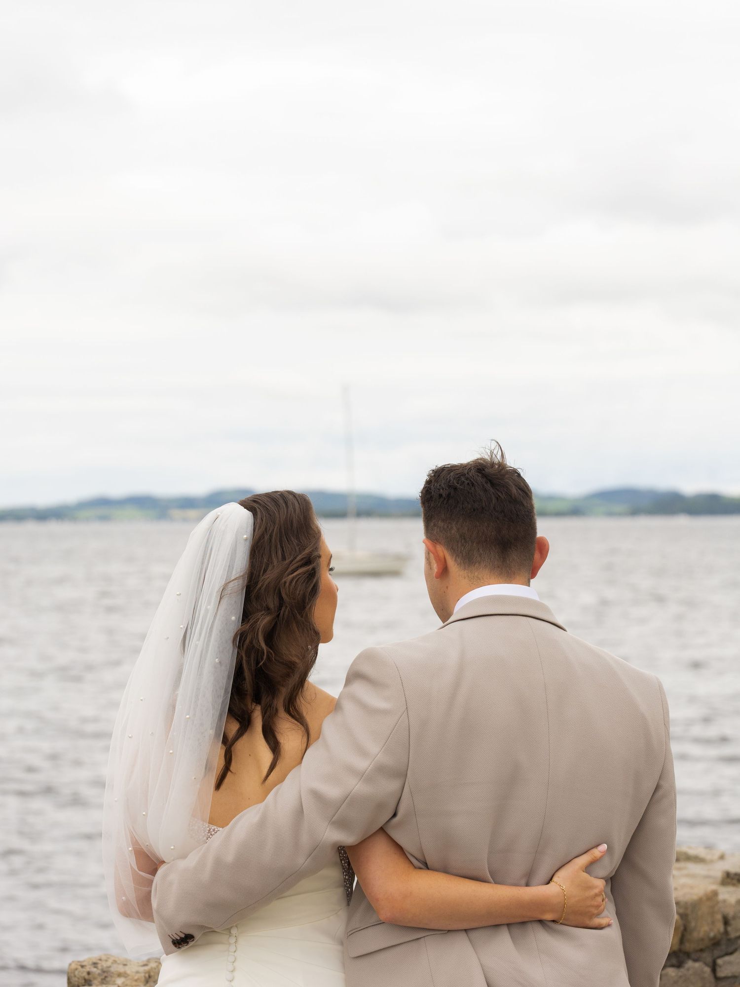 Newlywed couple embracing while looking out at the ocean from a rocky beach during their wedding photoshoot.