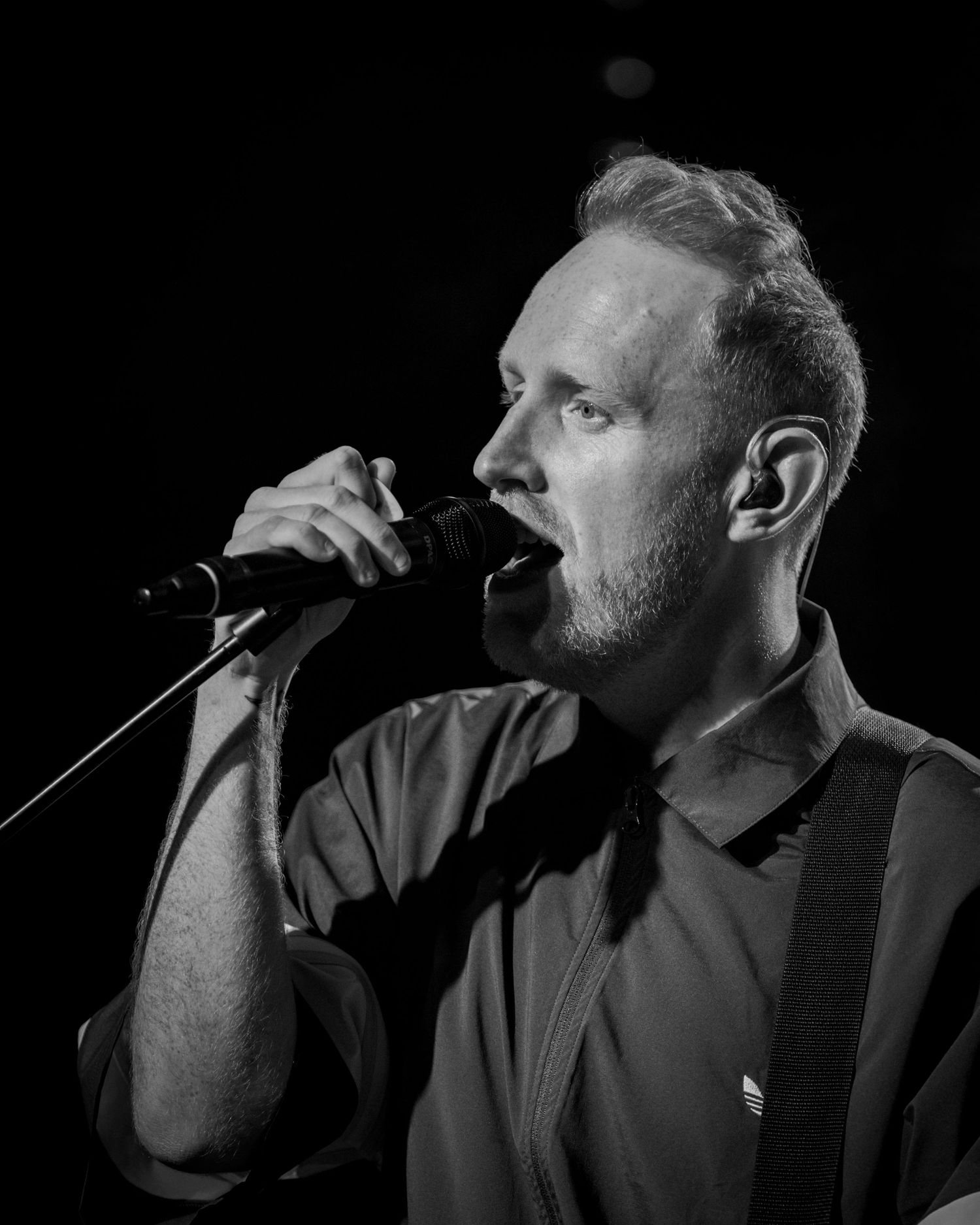 Black and white profile shot of performer singing into microphone on dark stage.