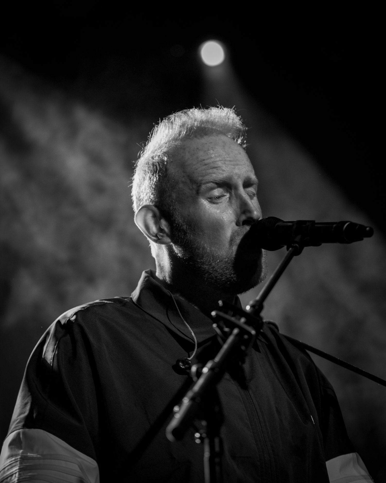 Black and white portrait of performer singing into microphone on stage with dramatic spotlight.