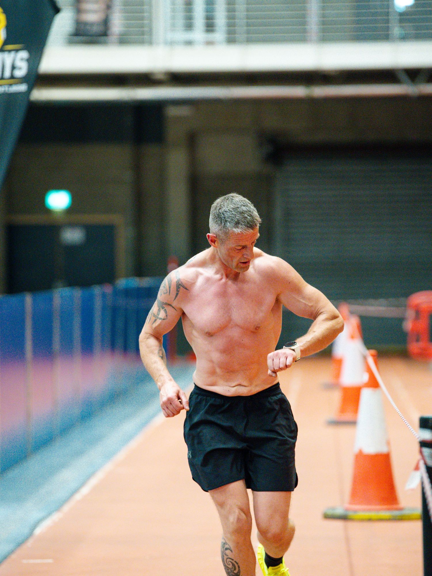Shirtless athlete running on a track with orange cones marking the course during training.