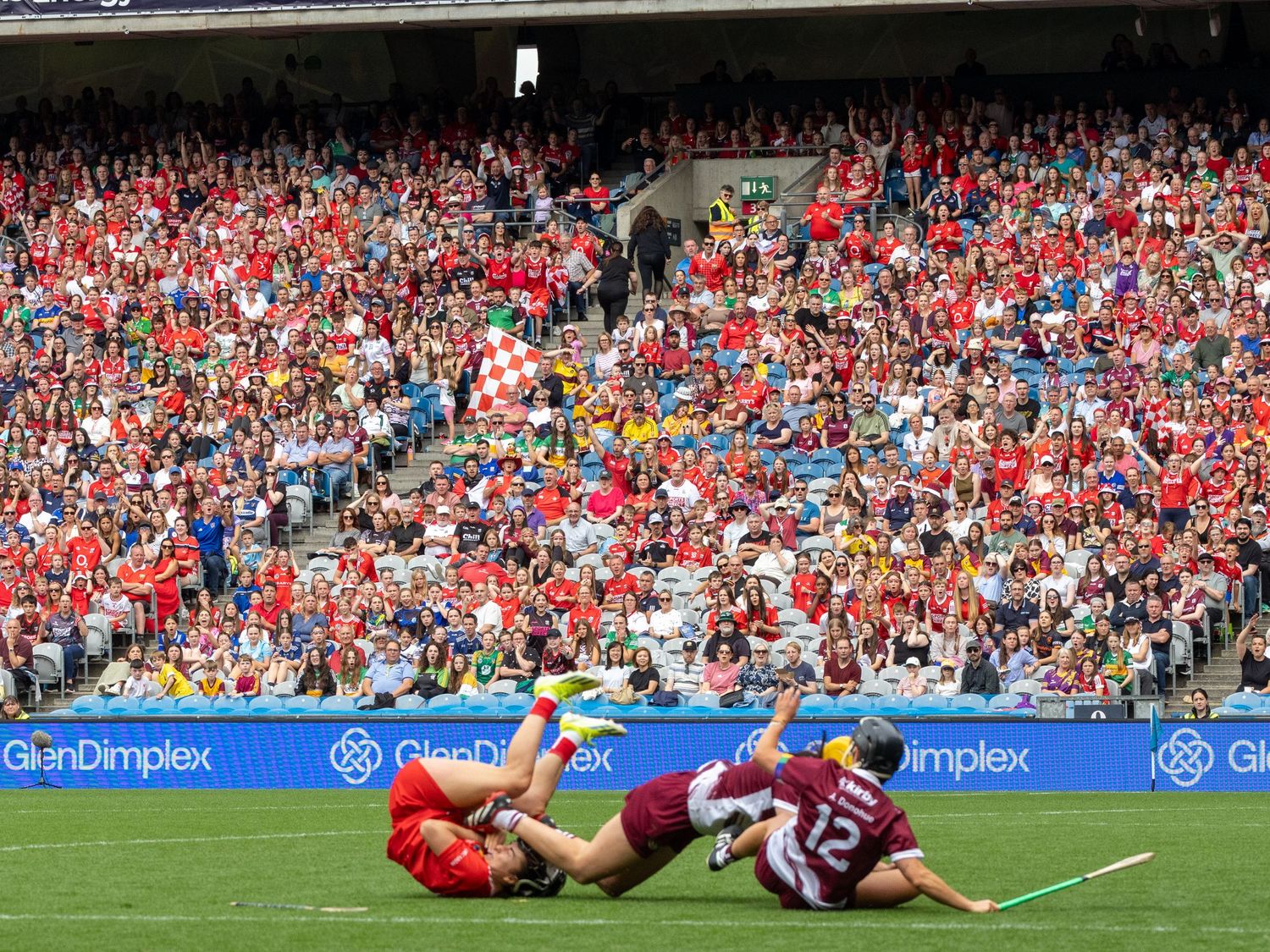 Two hurling players collide during a match as a packed crowd of red-clad supporters watch from the stadium stands.
