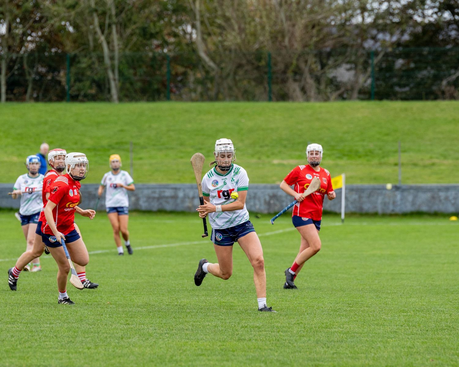 Players compete in a camogie match on a green field with players in red and white jerseys running with hurls.