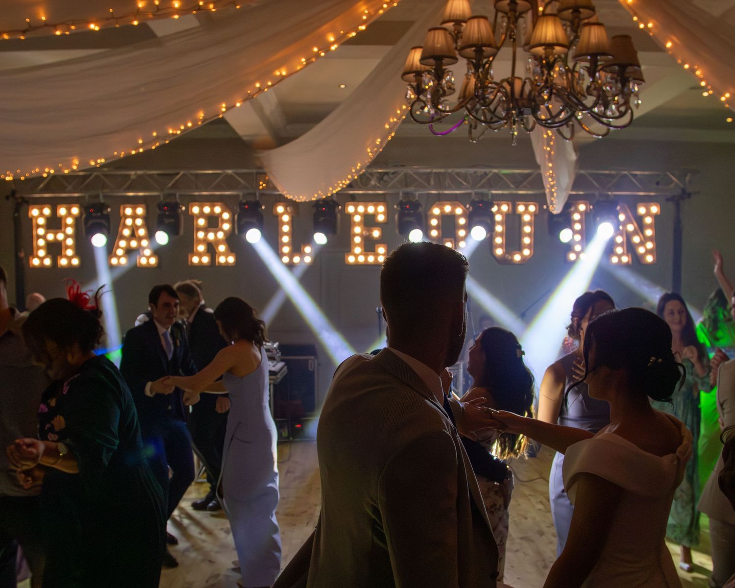 Silhouettes of people dancing at a wedding reception under twinkling string lights and white tent draping.
