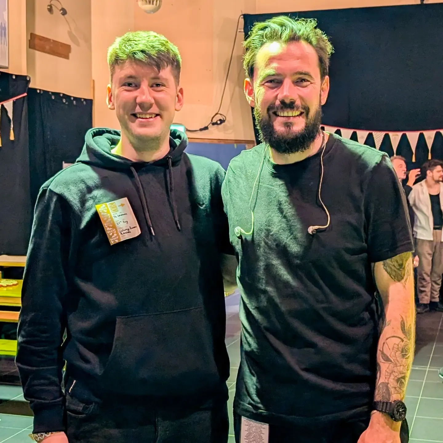 Two people in black clothing pose together at an event with a festive bunting banner visible in the background.