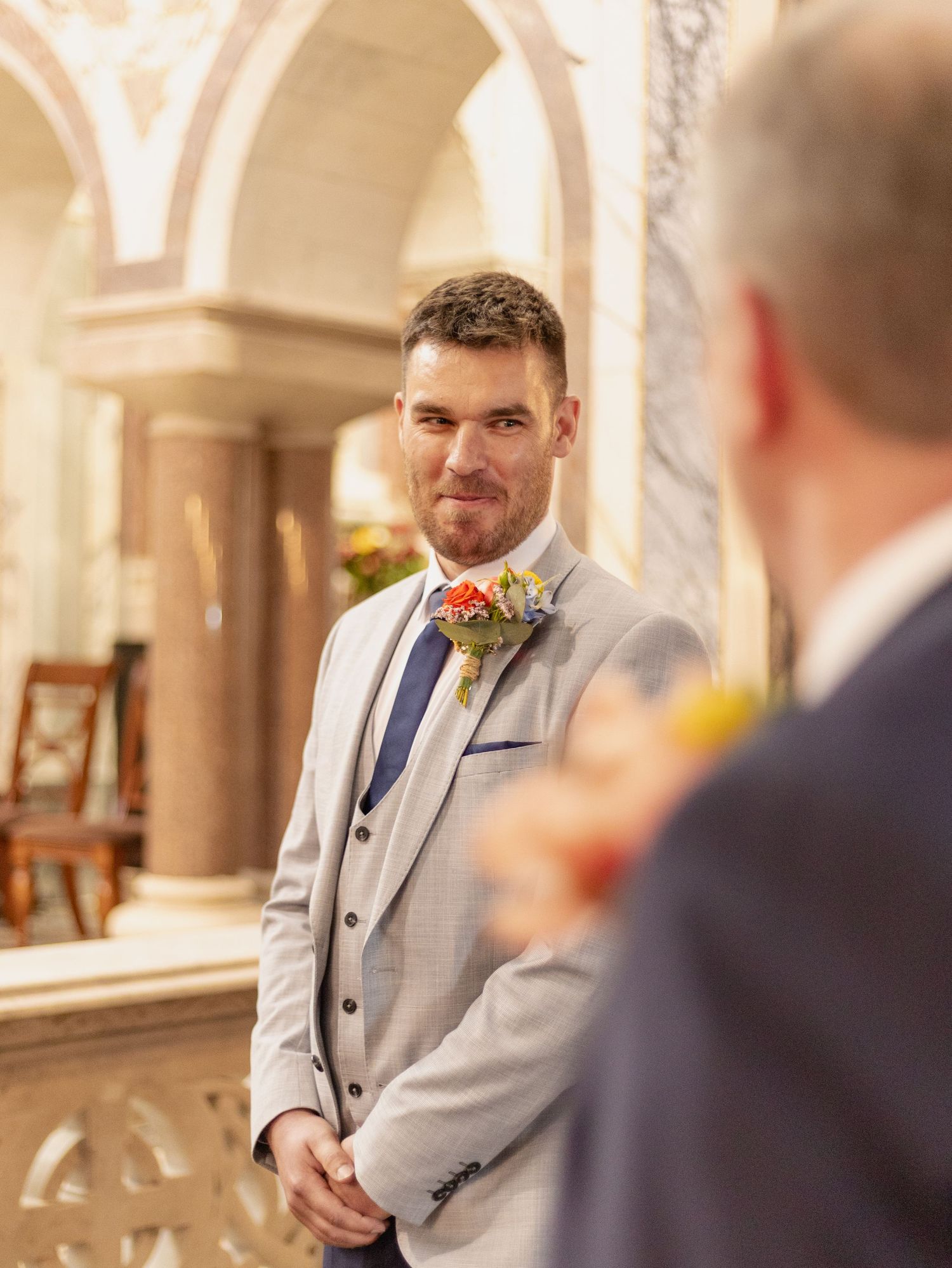 A person in a light gray suit jacket stands in an ornate room with classical architectural details.