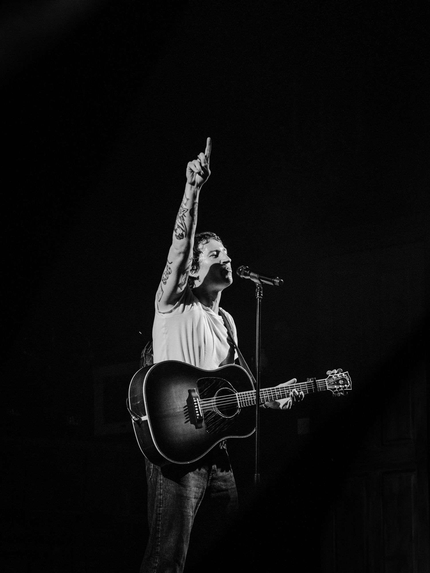 Black and white concert photo of acoustic guitarist pointing upward while performing on stage.