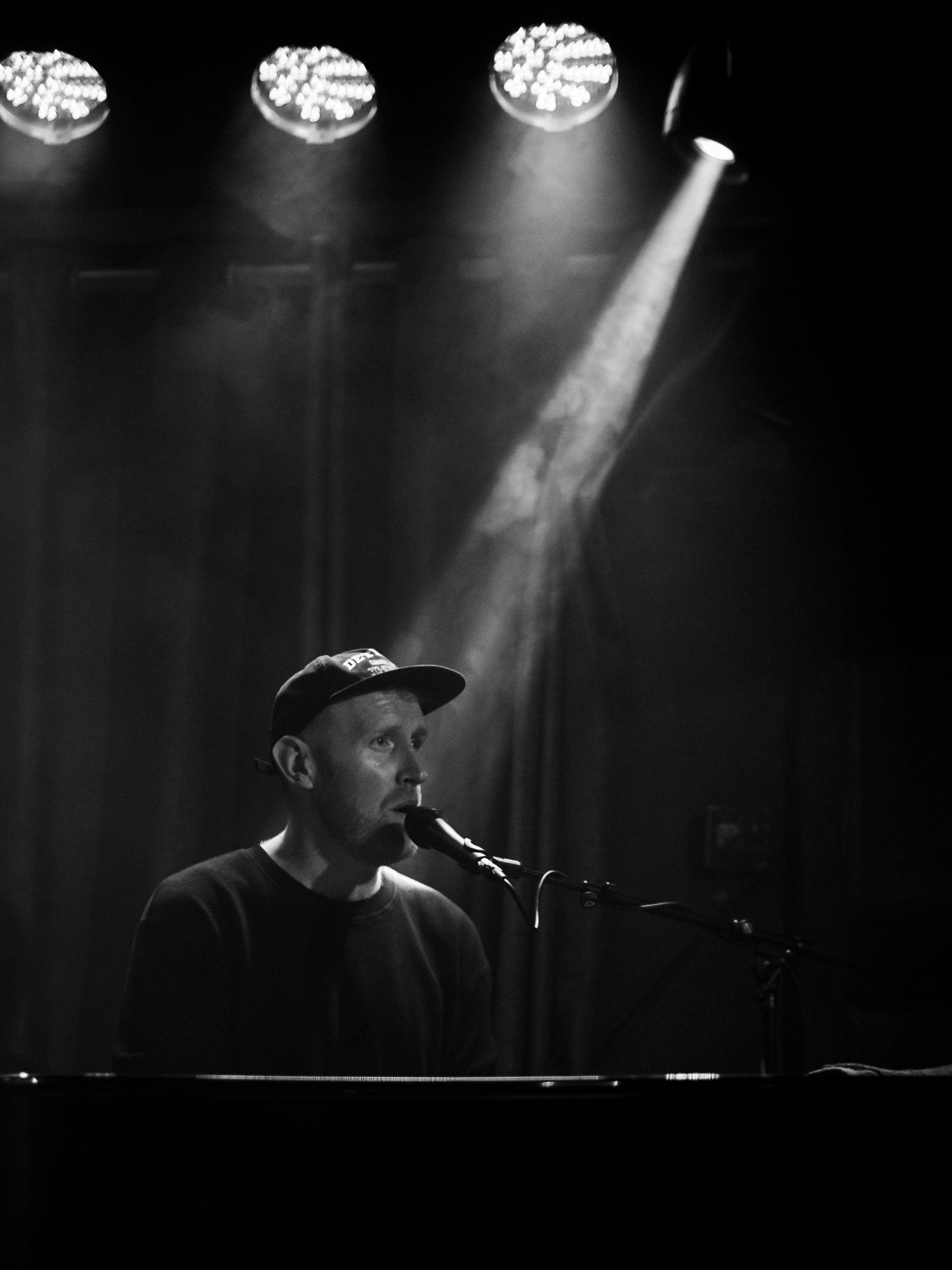 A performer plays piano and sings on a dimly lit stage with dramatic spotlights illuminating the scene in black and white.
