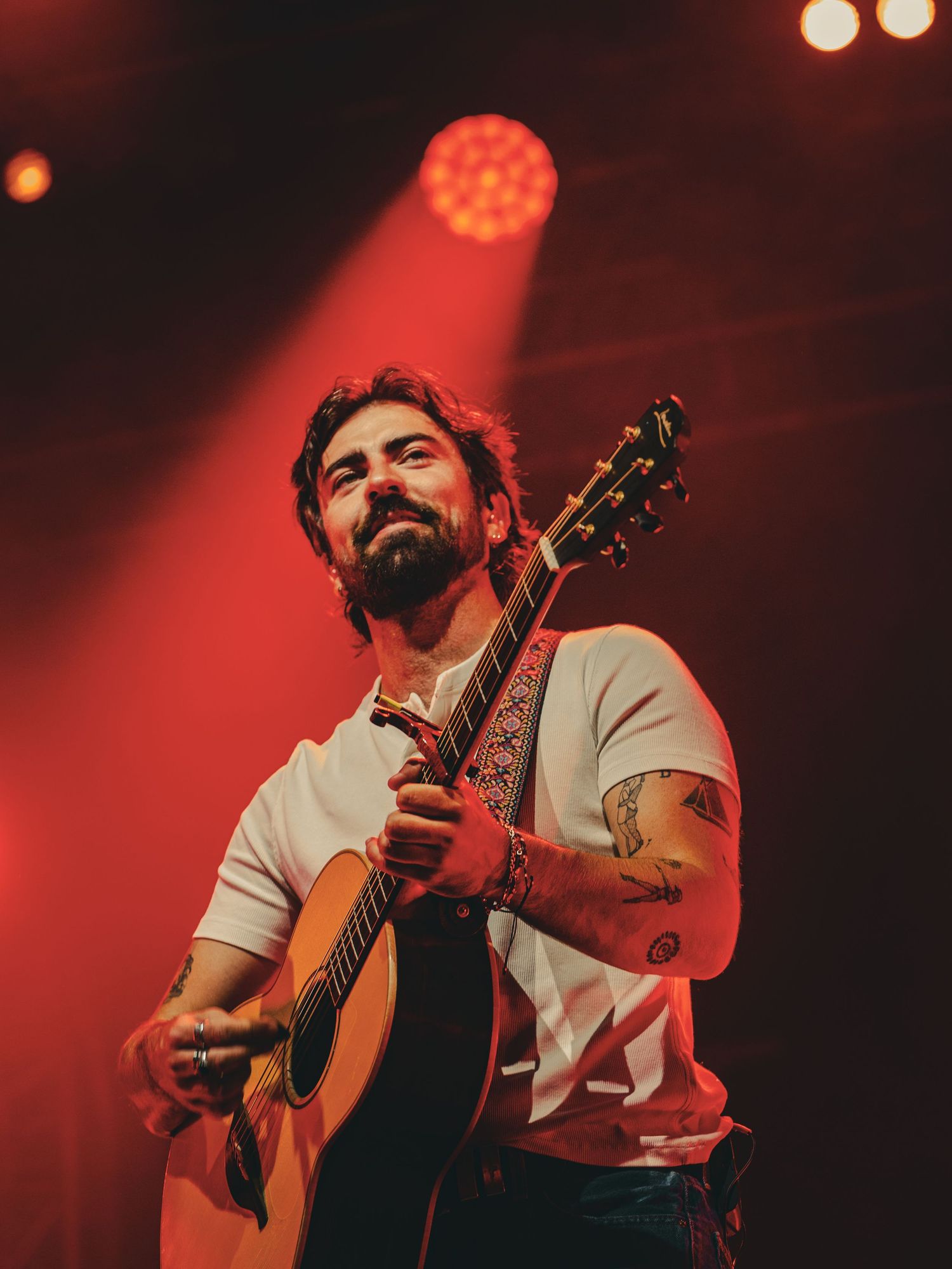 Musician performs on acoustic guitar under dramatic red stage lighting at a concert venue.