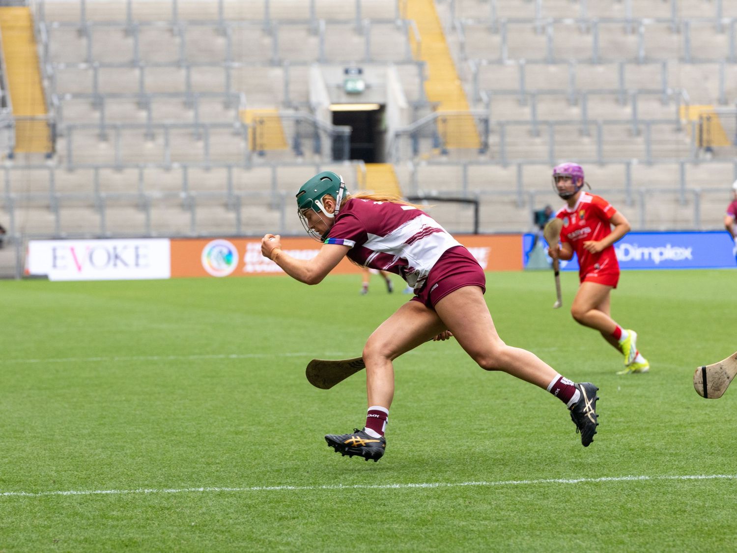 A hurling player in maroon jersey dives to strike the ball during a match at a stadium.