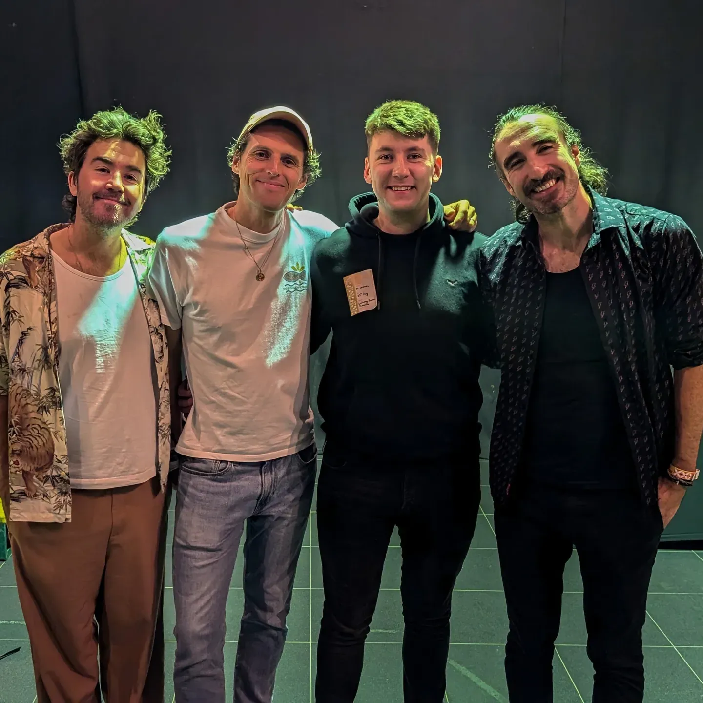 Group of performers wearing casual attire and green accessories pose together on a stage.