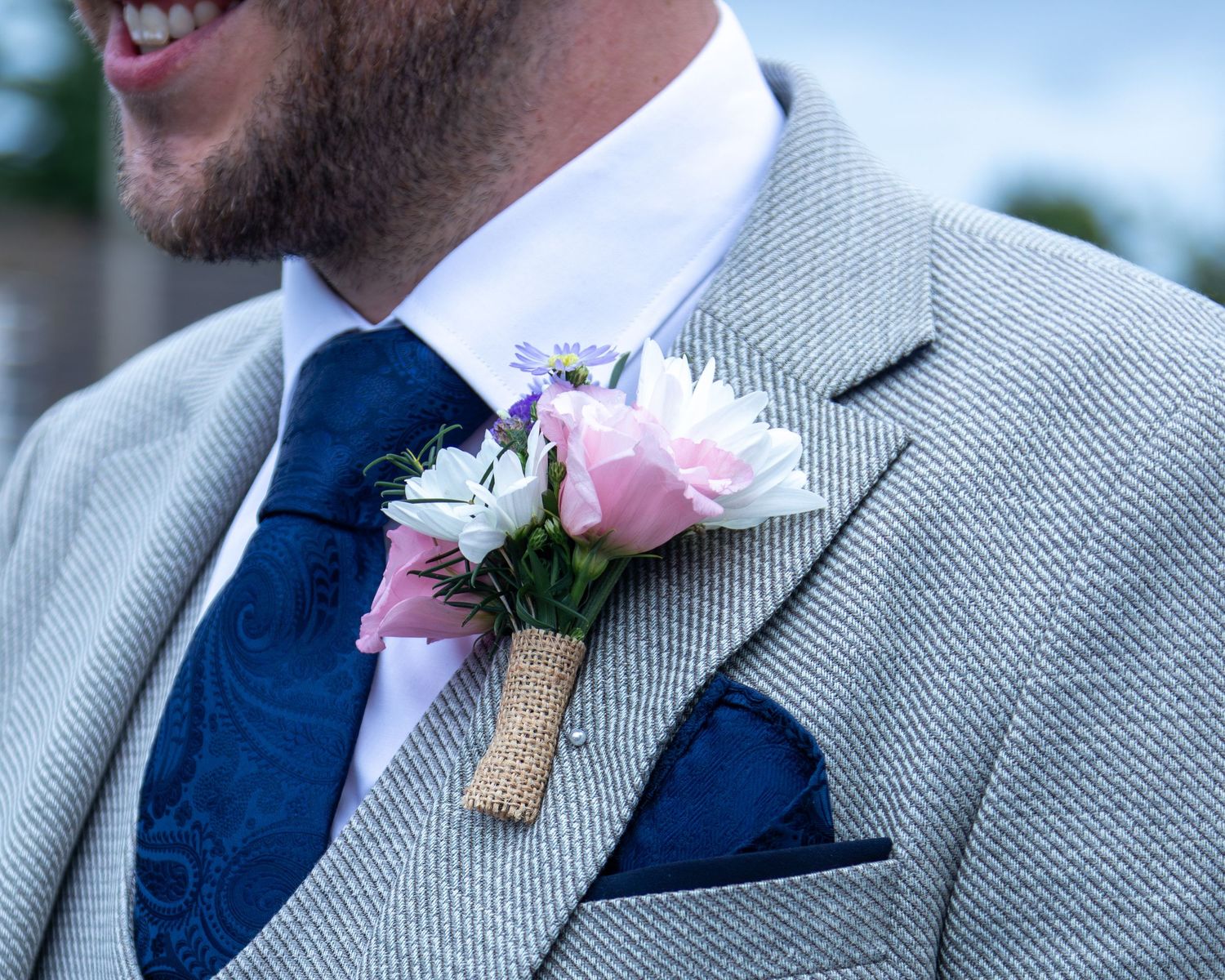 Close-up detail of wedding attire showing light gray suit jacket with navy tie and pink rose boutonniere.