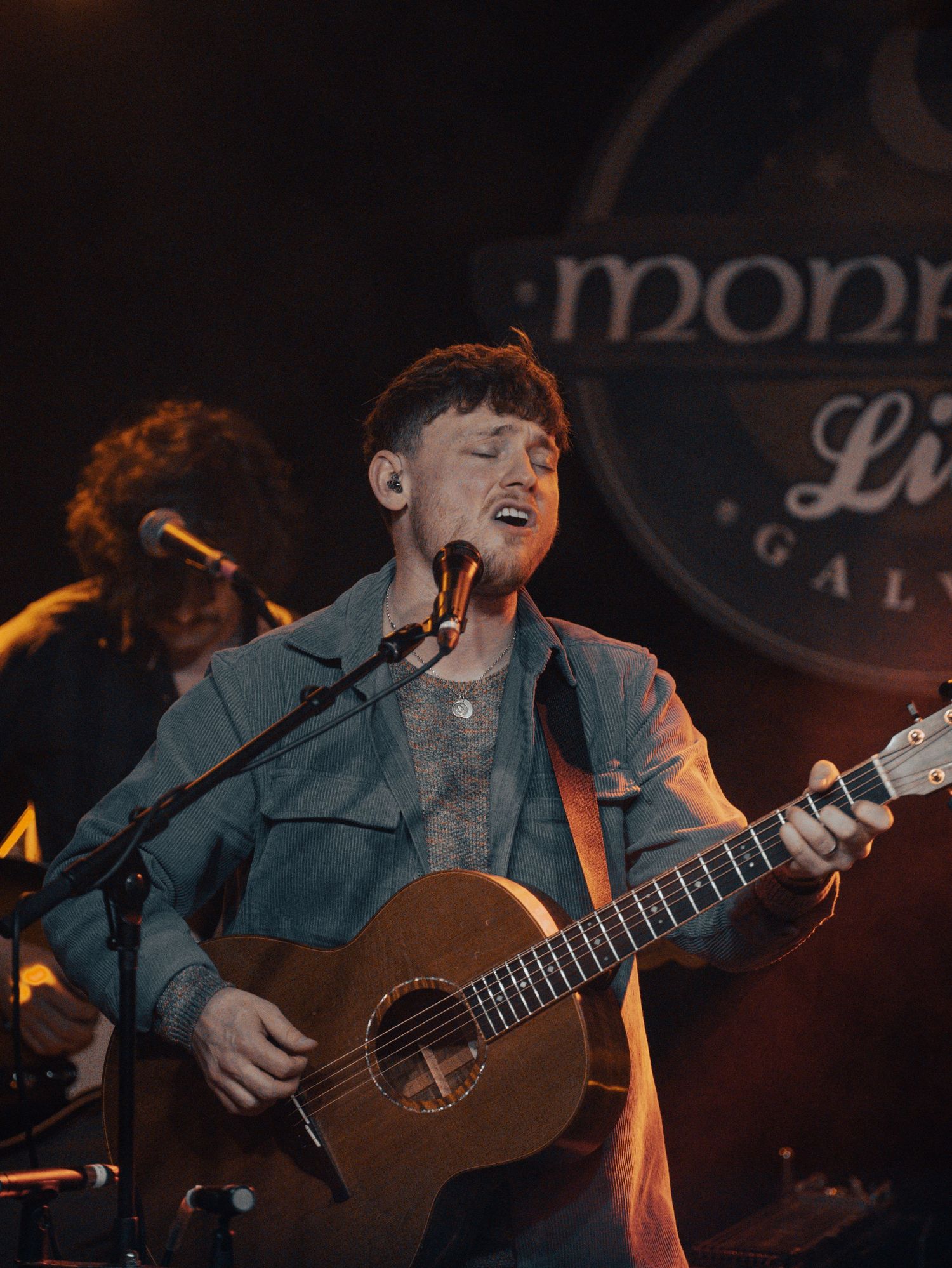 Musician performing with acoustic guitar under warm lighting with venue signage visible in background.