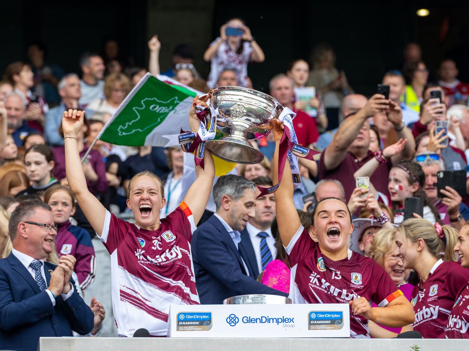 Excited sports fans in maroon jerseys celebrate and cheer during a match while holding up a trophy.