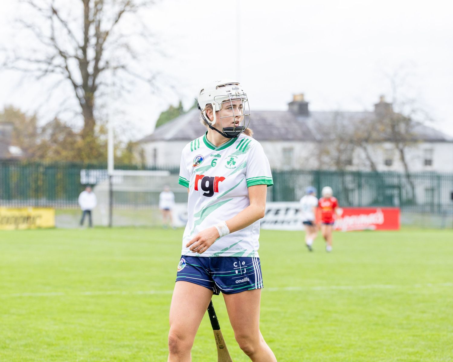 Camogie players in white and green jerseys compete during a match on a grass field.