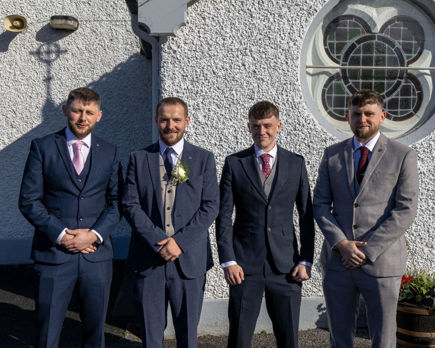 Groomsmen in dark suits lined up against white church wall.