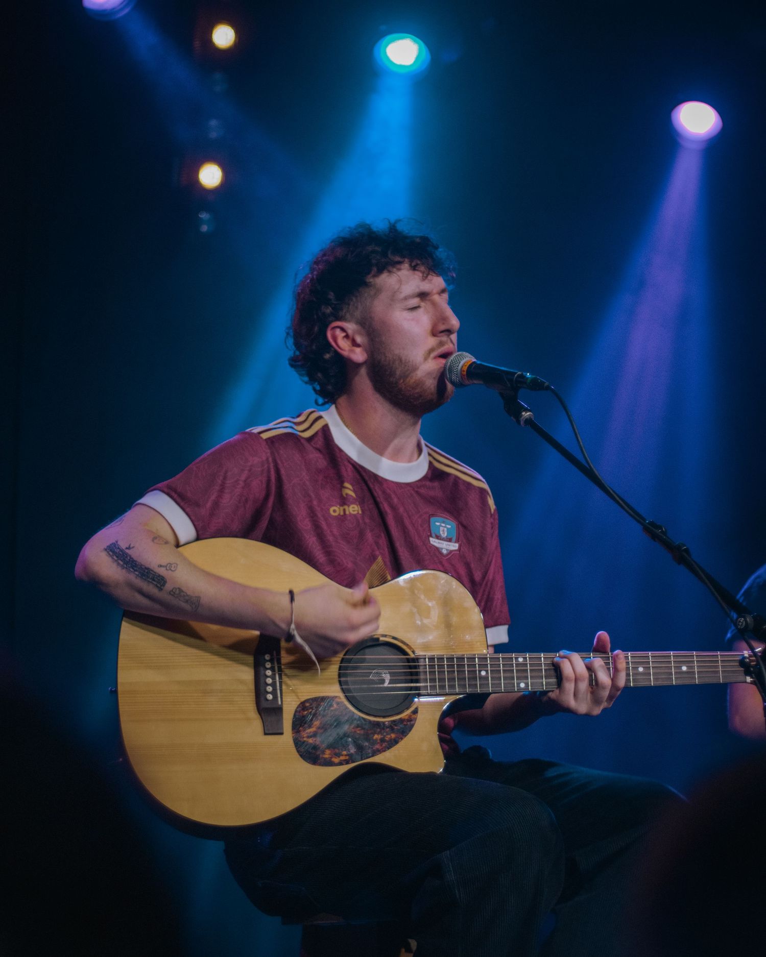 A musician in a maroon jersey plays acoustic guitar while singing into a microphone under colorful stage lights.