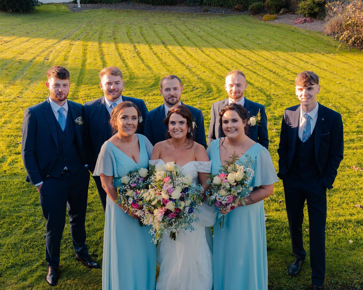 Wedding party in light blue dresses and navy suits poses in sunny garden.