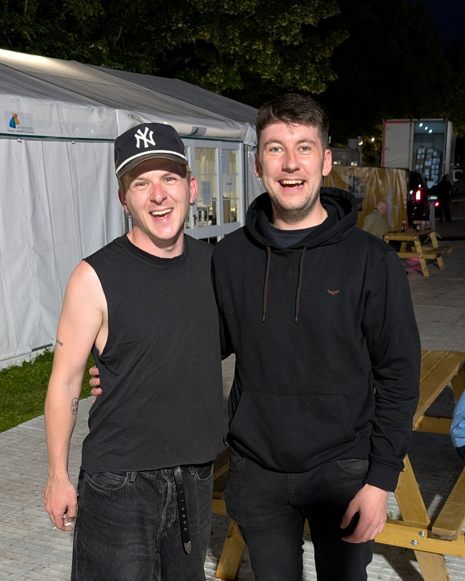 Two people wearing black clothing stand together smiling outdoors at night near picnic tables.