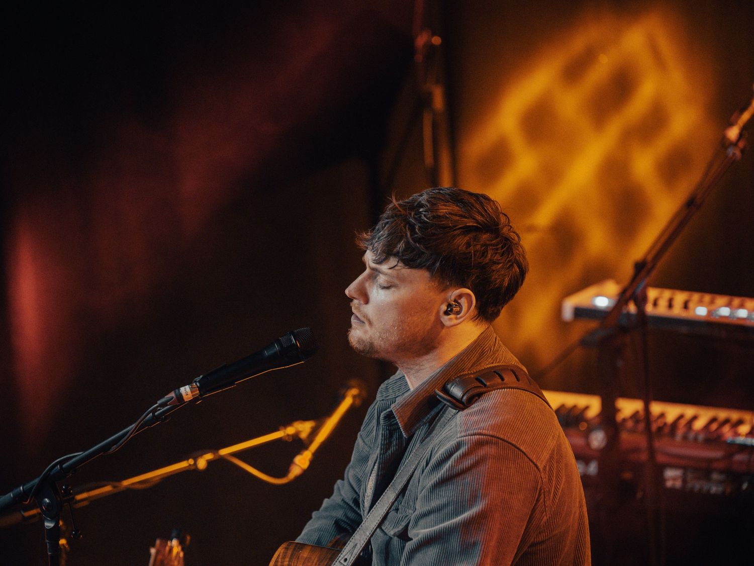 Concert performer on stage with warm orange lighting and musical equipment in background.