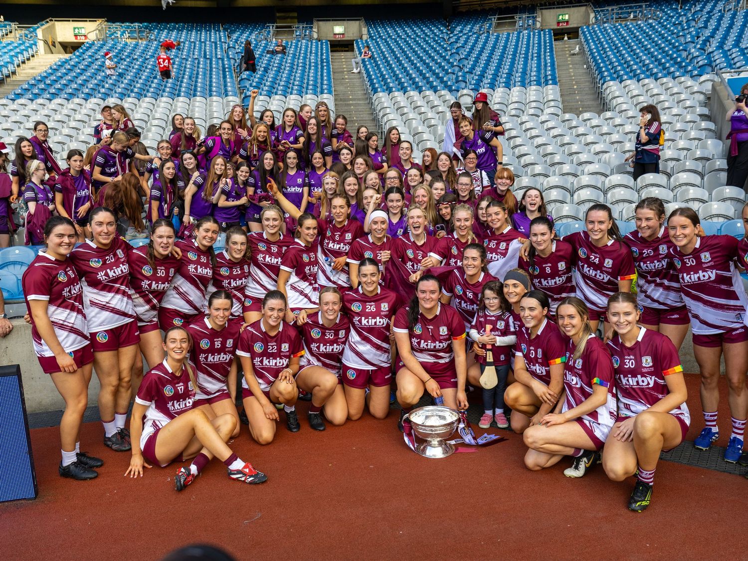 Large group of athletes in maroon uniforms pose together on stadium field after victory celebration.