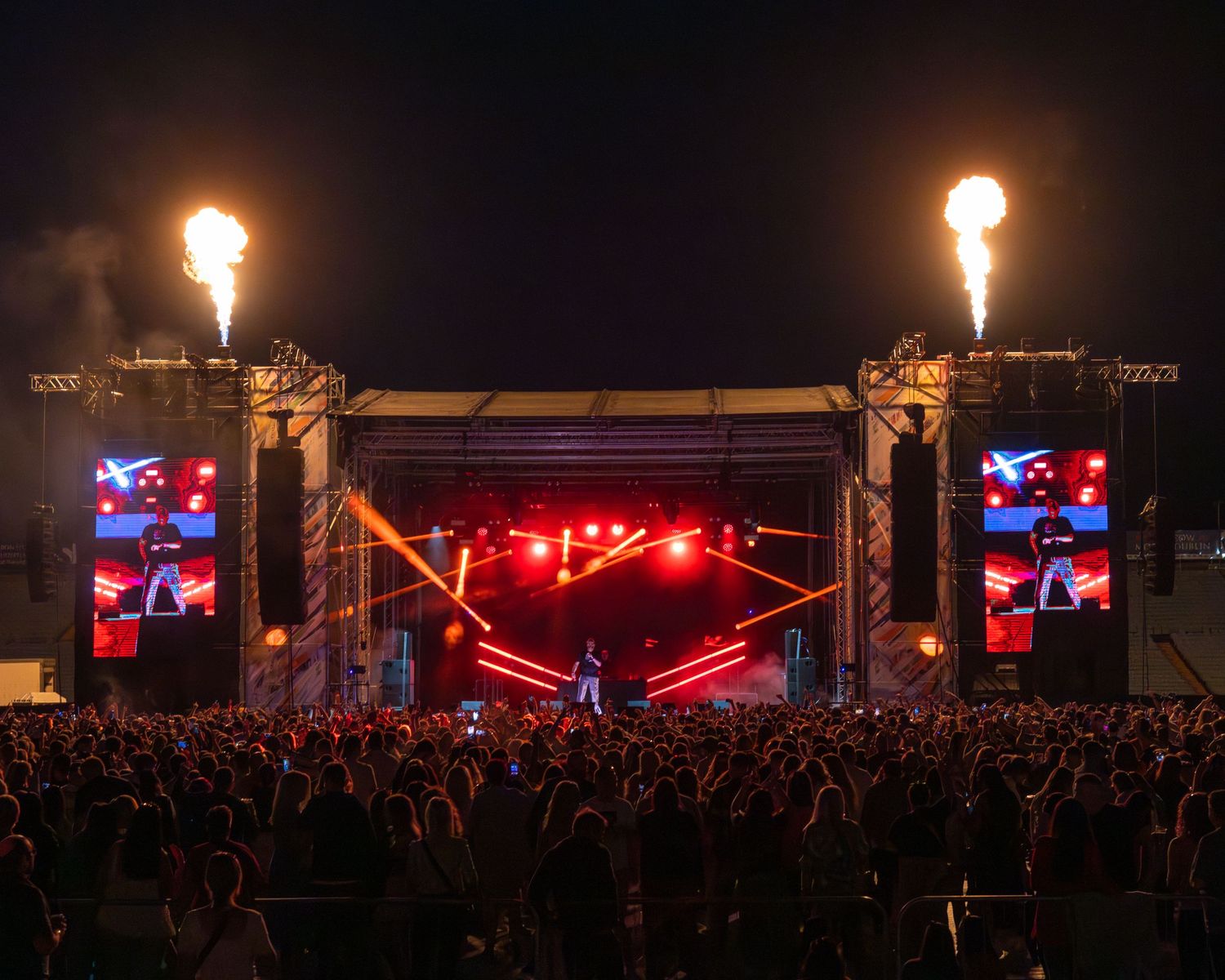 Concert stage with pyrotechnic flames and large LED screens illuminating a crowd at an outdoor music festival.