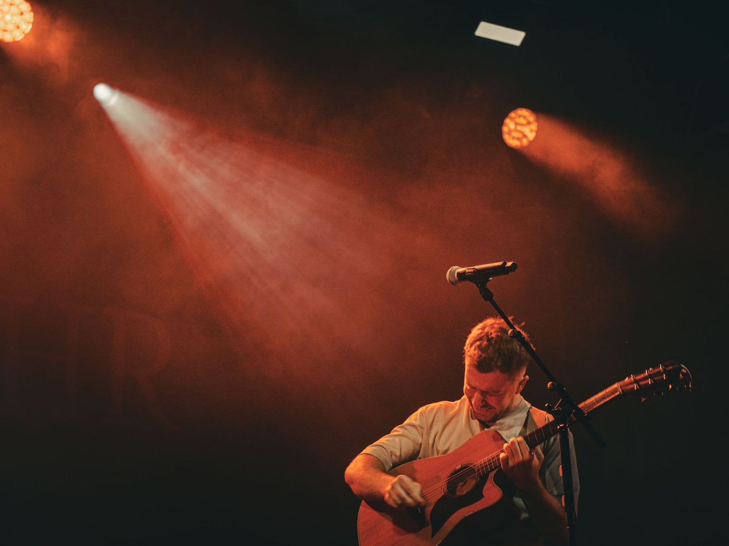 Musician plays acoustic guitar under warm orange spotlights on dark stage.