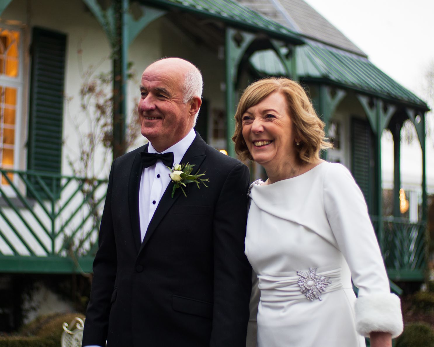 A couple in formal wedding attire share a joyful moment together outdoors near a green-trimmed building.