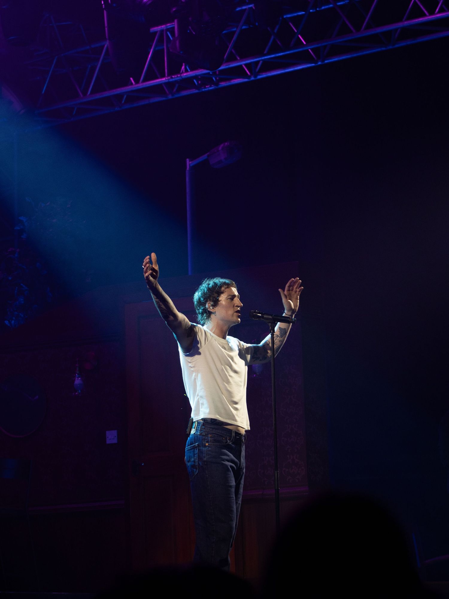 A performer in a white shirt and jeans stands on stage with raised arms under dramatic blue lighting during a concert.