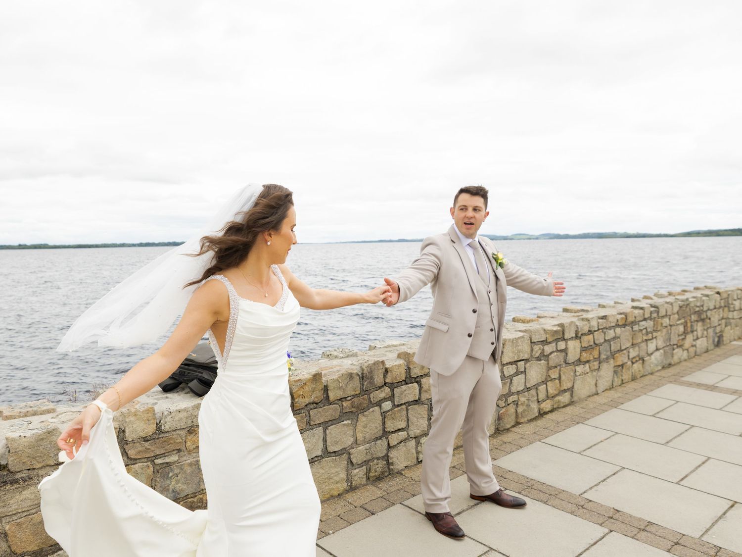 Wedding couple walks hand in hand along a stone wall overlooking a serene waterfront vista.