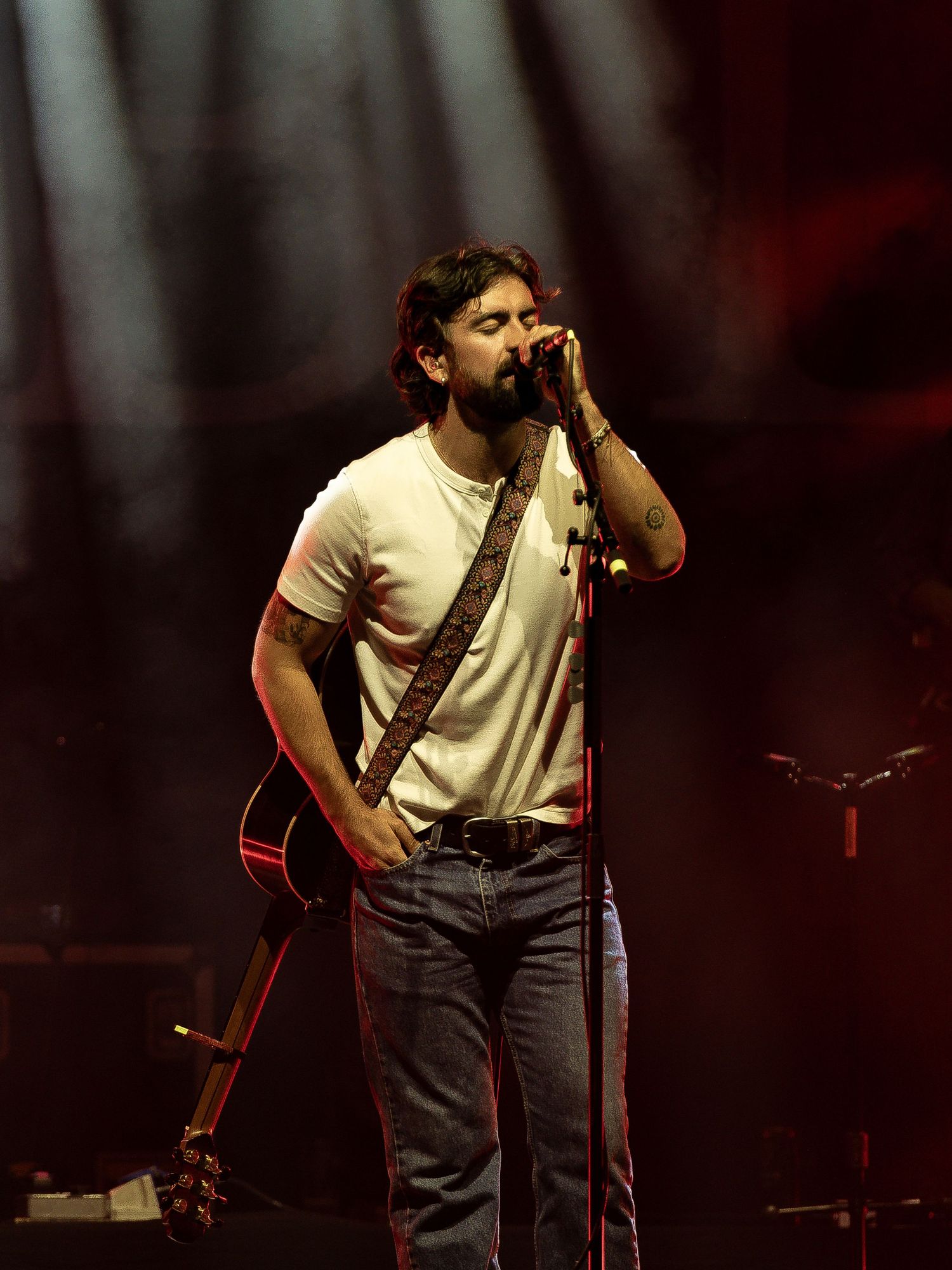 Concert performer singing into microphone while holding acoustic guitar on dimly lit stage.