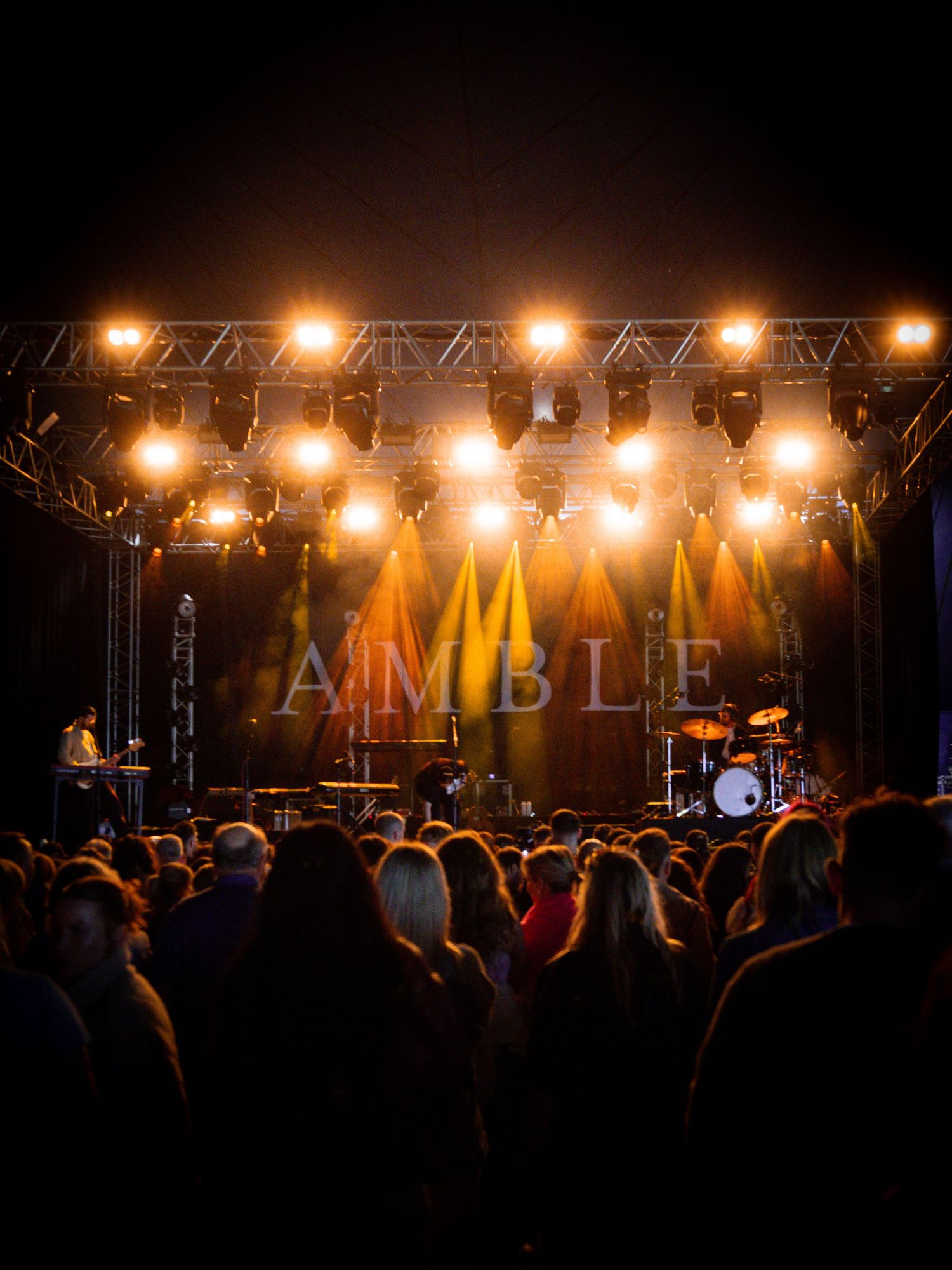 Concert stage illuminated with warm orange lights and truss rigging casting dramatic shadows over a nighttime crowd.