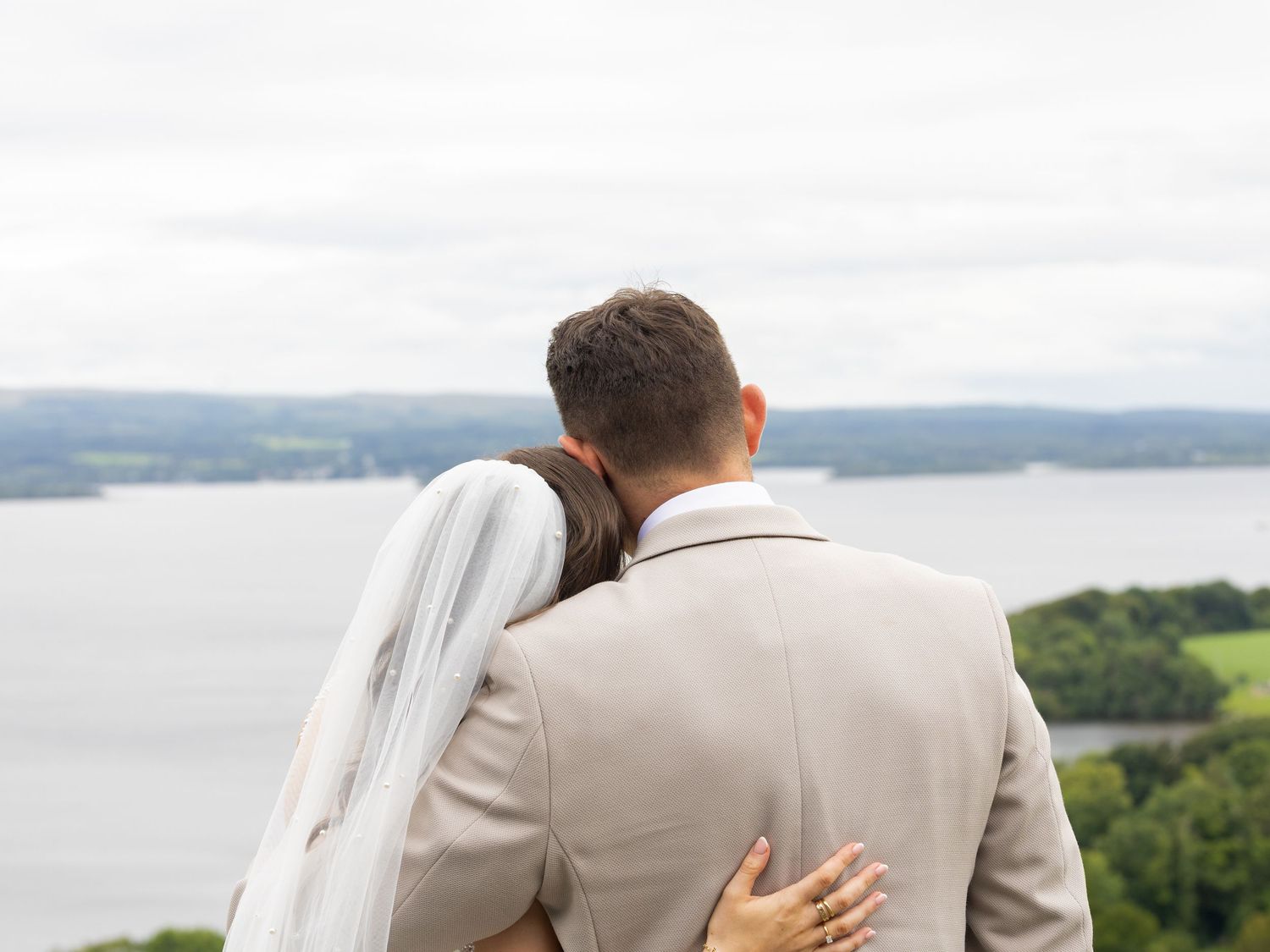 A couple in wedding attire embraces while overlooking a scenic lake view from an elevated vantage point.
