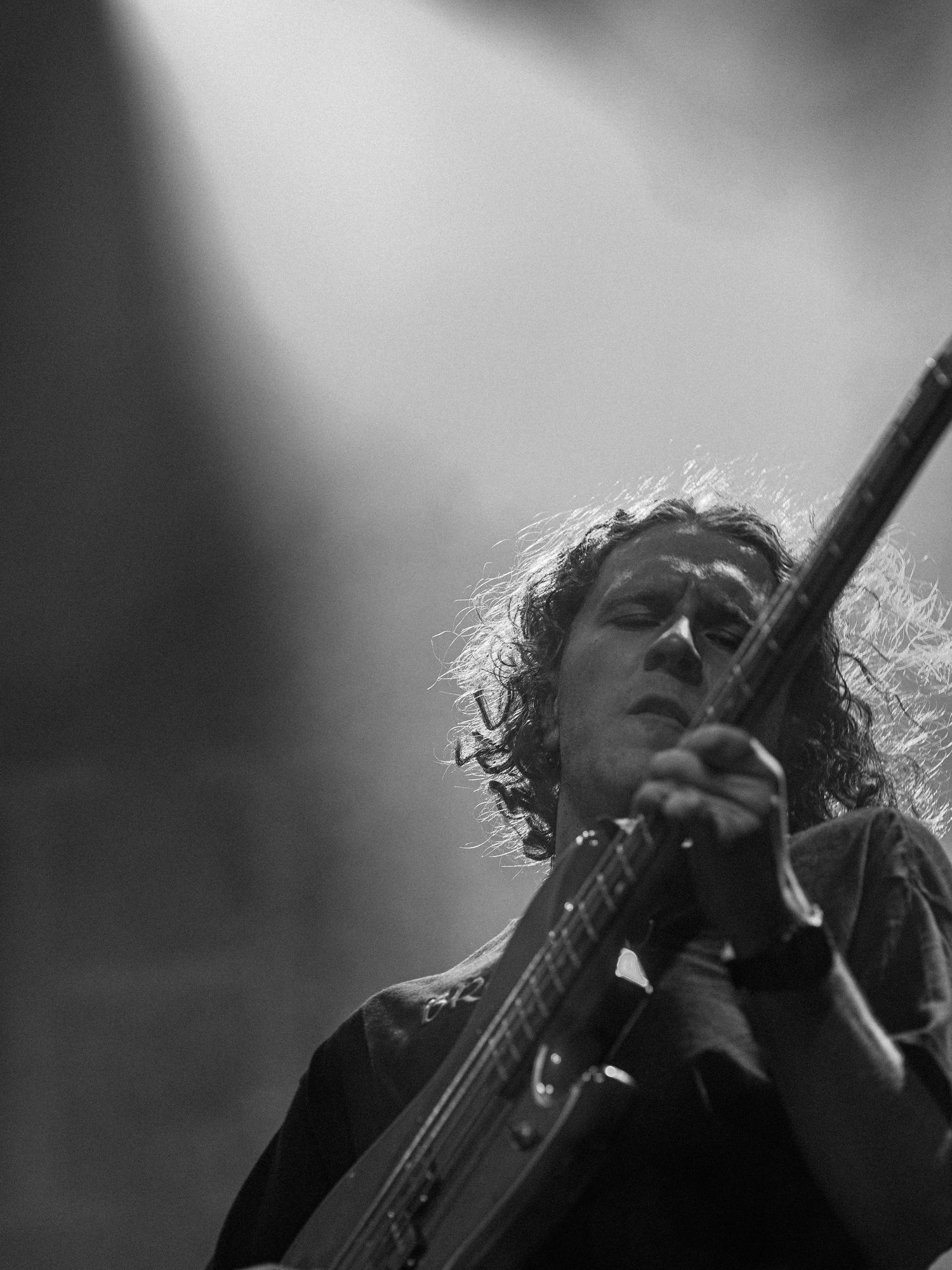 Musician with curly hair plays guitar passionately in black and white stage lighting.