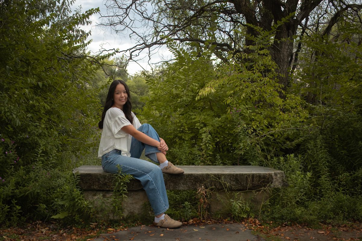 A person sits on a wooden bench surrounded by lush green foliage in a peaceful outdoor setting.