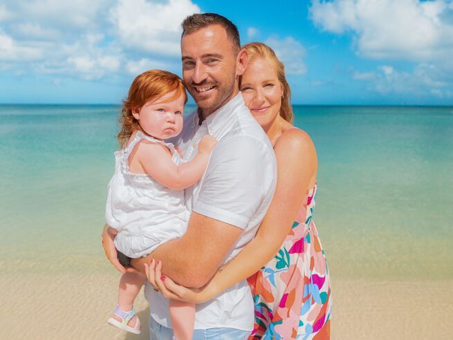 Happy family of three posing on a tropical beach with turquoise water, parents holding a baby girl.