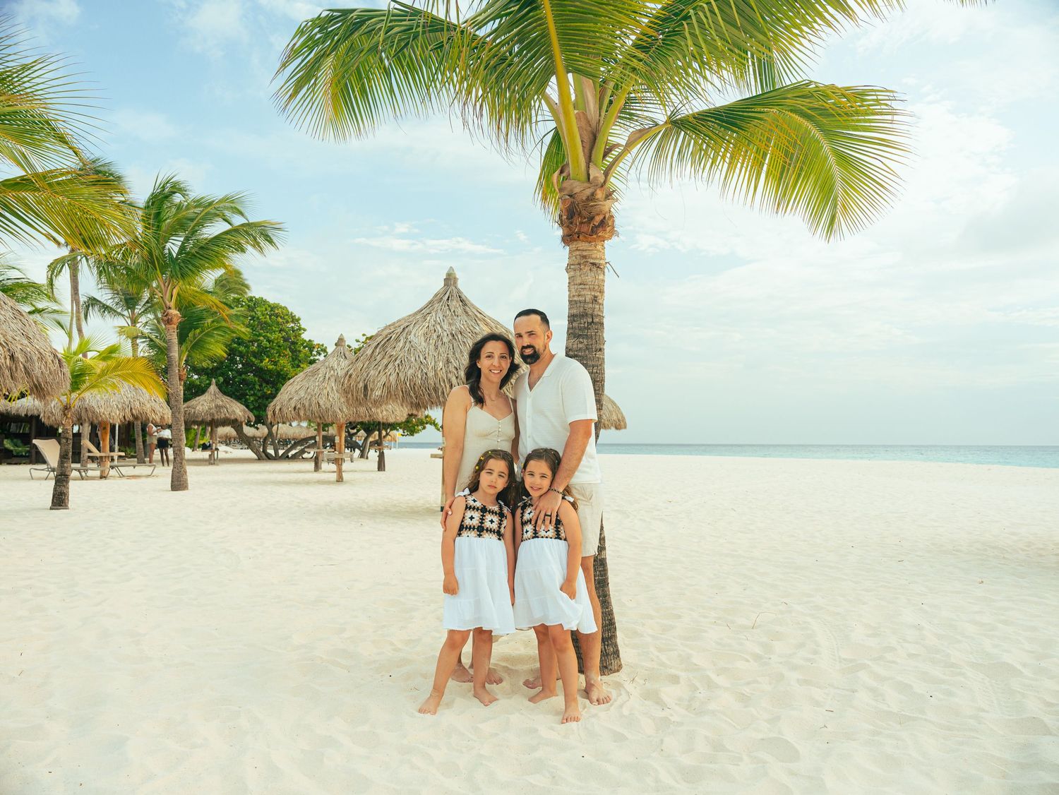Happy family of four posing under a palm tree on a tropical white sand beach.