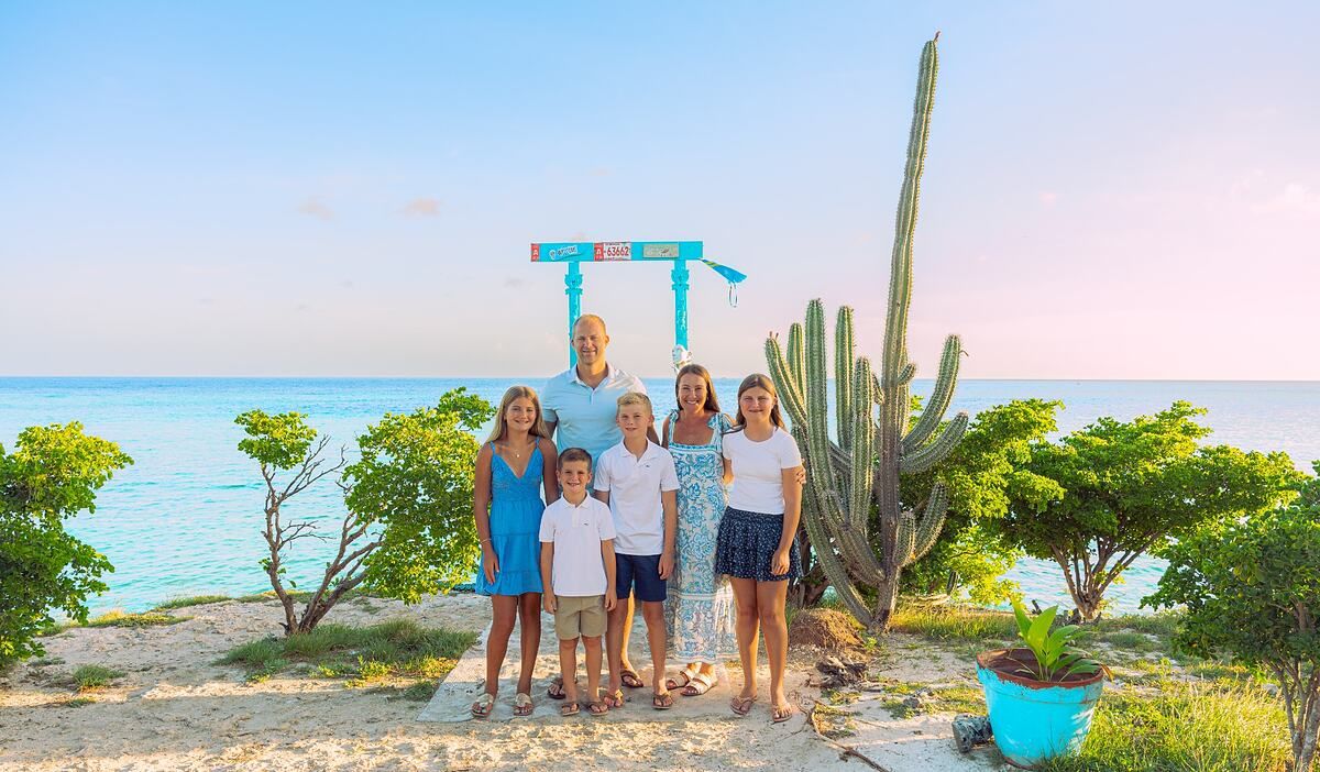 Family of six poses on a tropical beach with cactus and turquoise water at sunset.