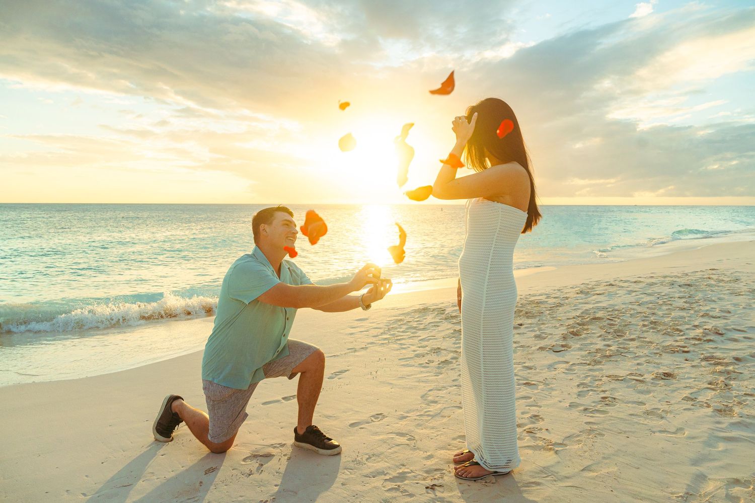 Man proposing on one knee to a woman on a white sandy beach at sunset with rose petals in the air.