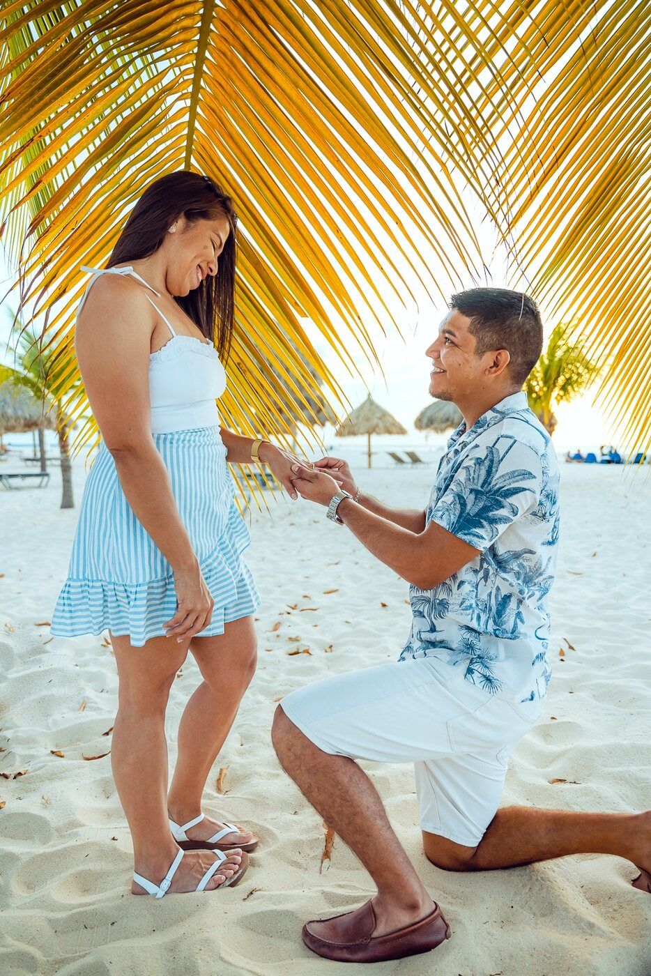 Man proposes to woman on one knee on a tropical beach under palm trees, romantic marriage proposal scene.