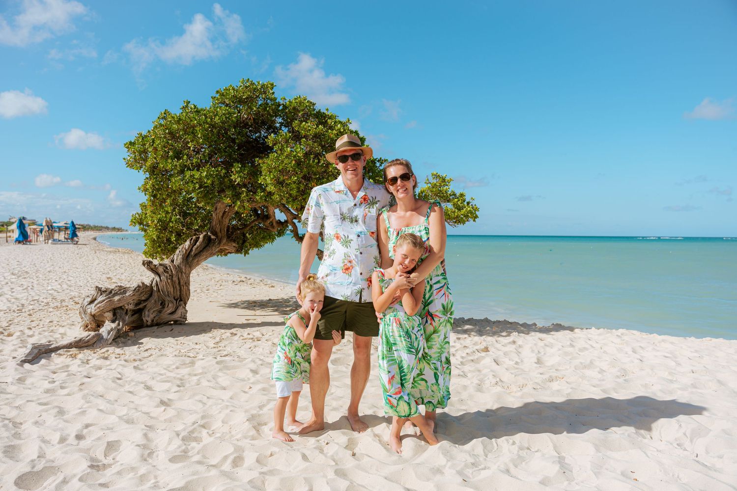 Family of four posing on white sandy Aruba beach near iconic divi tree with turquoise ocean backdrop.