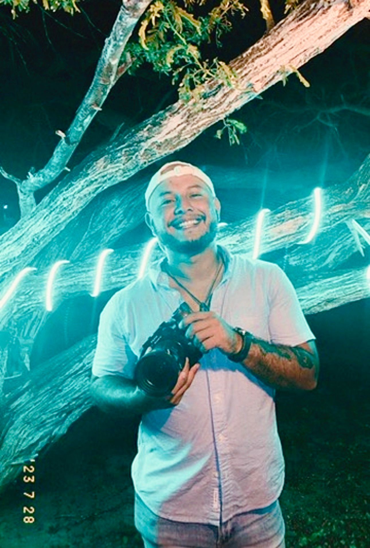 Smiling tattooed photographer holding a camera under a tree illuminated by teal light at night.
