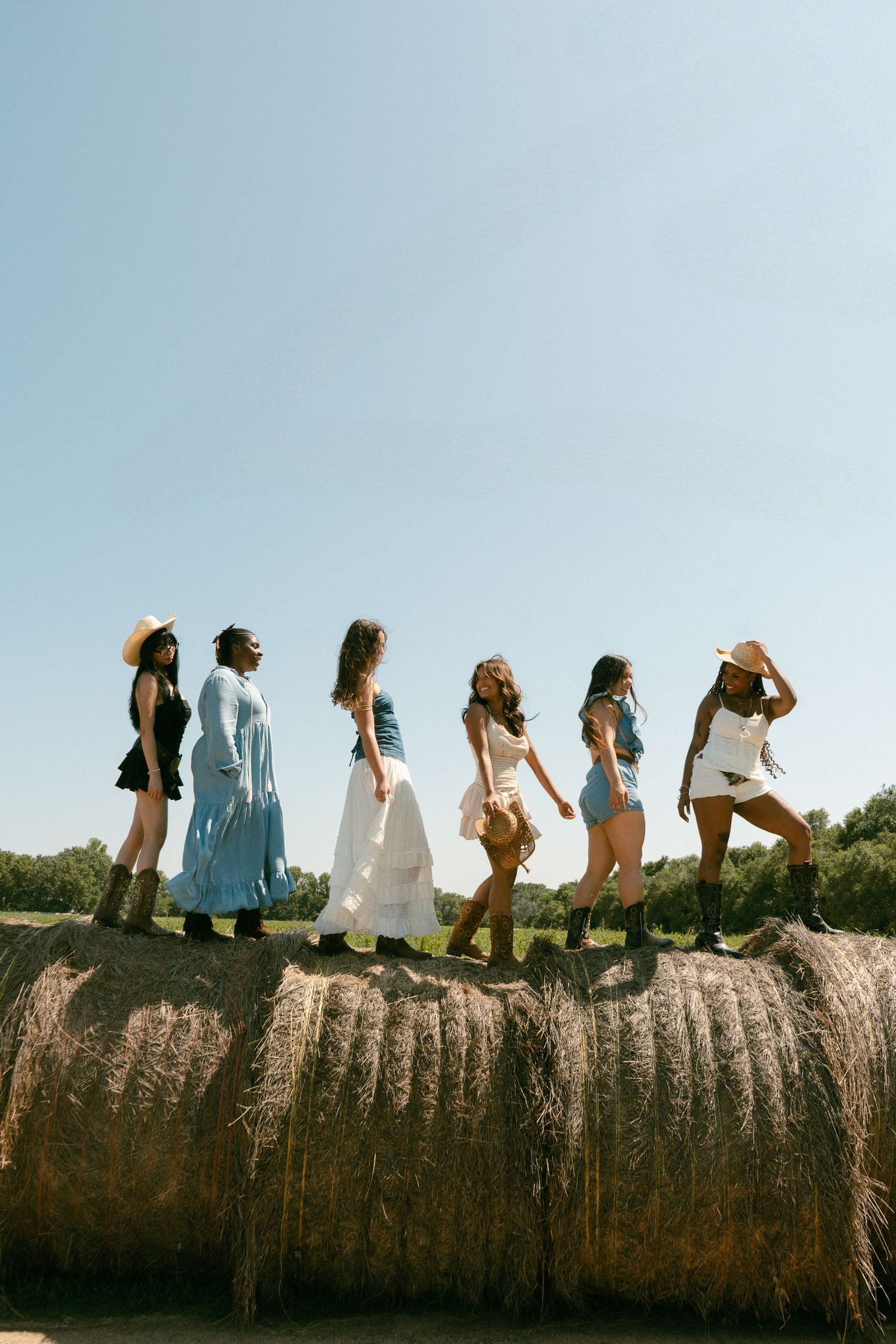 Five diverse women in western outfits standing on a large hay bale in a sunny field.
