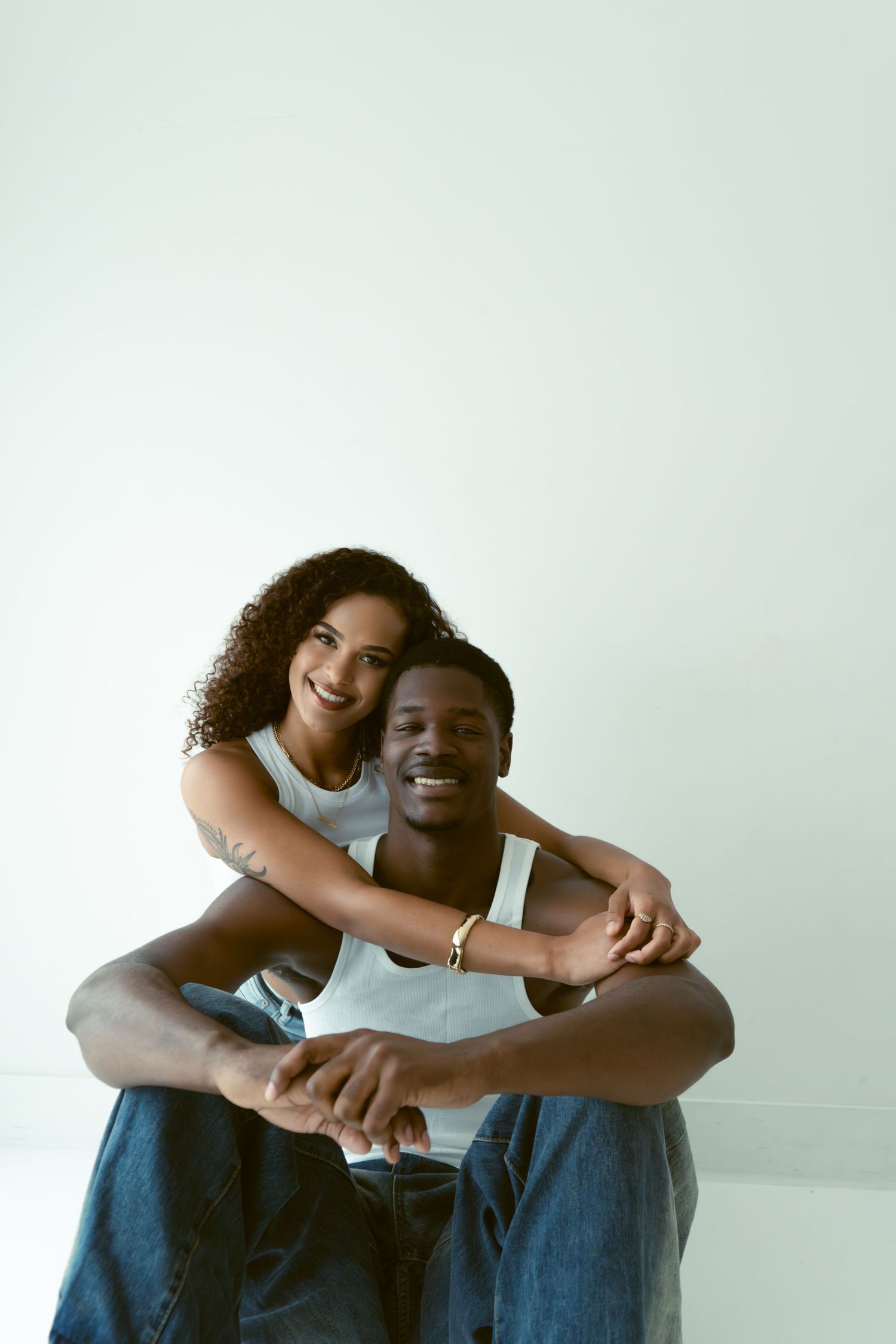 Happy couple embracing against white wall, woman hugging man from behind, both smiling in jeans.