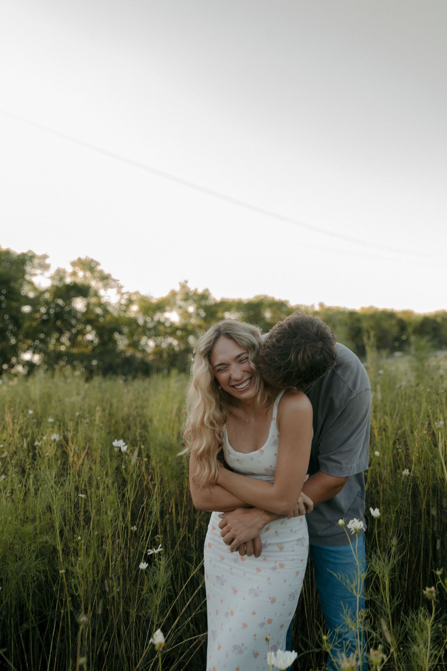 Happy blonde woman in floral dress laughing as man embraces her from behind in wildflower field.