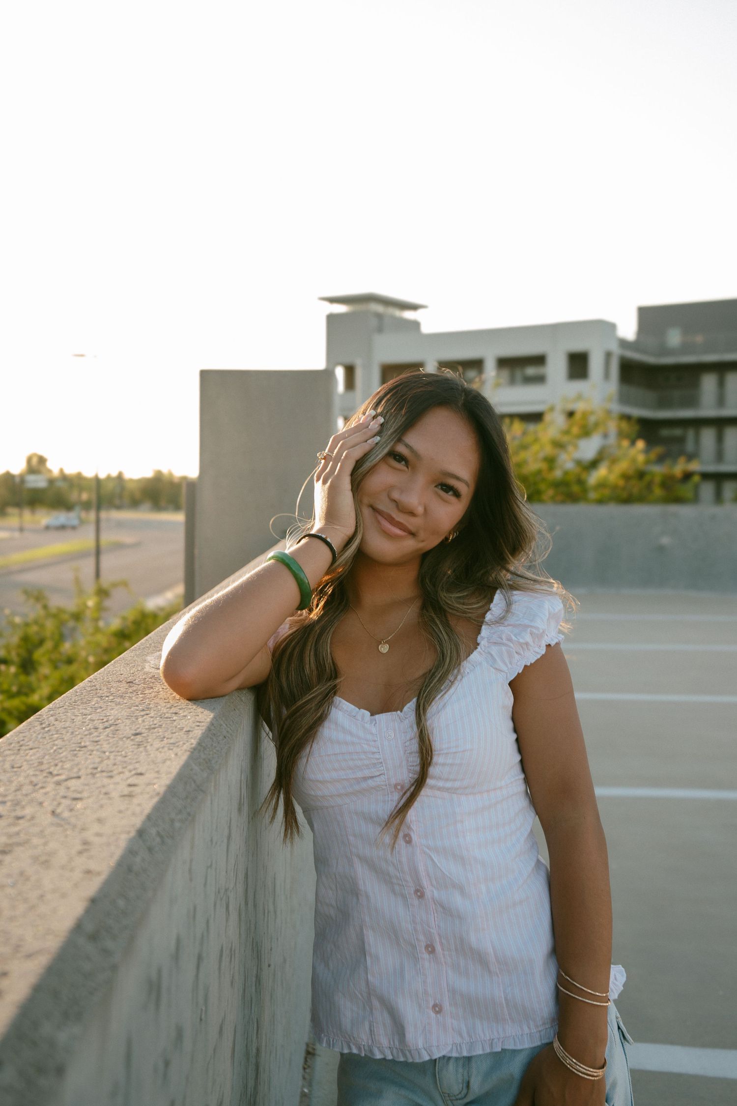 Young woman in white top leaning against concrete wall at golden hour rooftop parking lot.