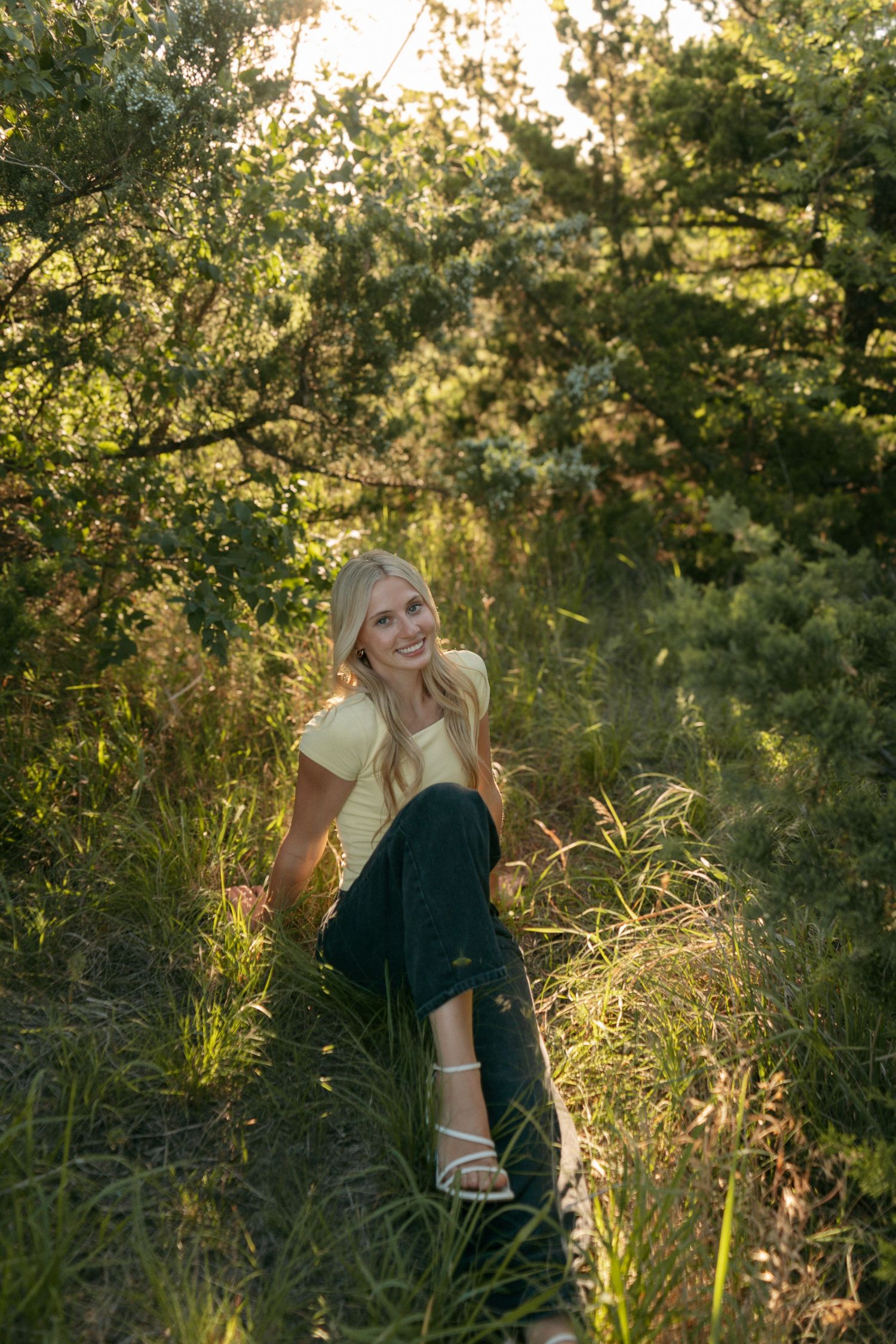 Smiling blonde woman sitting in lush green grass surrounded by trees, wearing yellow top and dark jeans in golden sunlight.