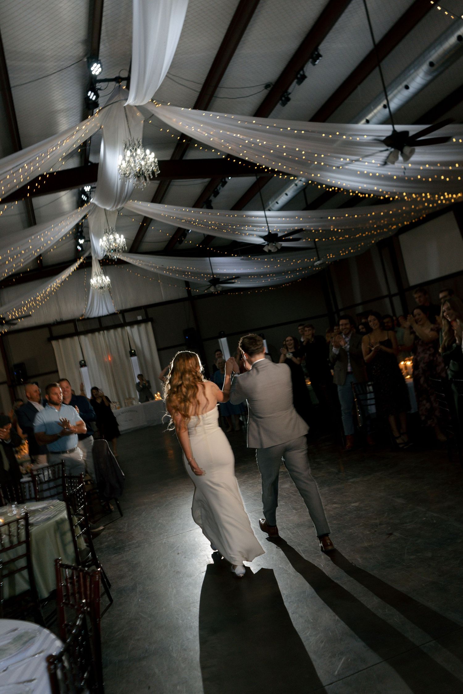 Bride and groom share first dance in elegantly decorated reception hall with fairy lights and white draping.