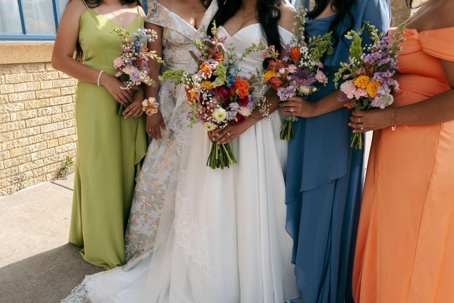 Bride in lace gown with bridesmaids in green, blue, and orange dresses holding colorful floral bouquets.