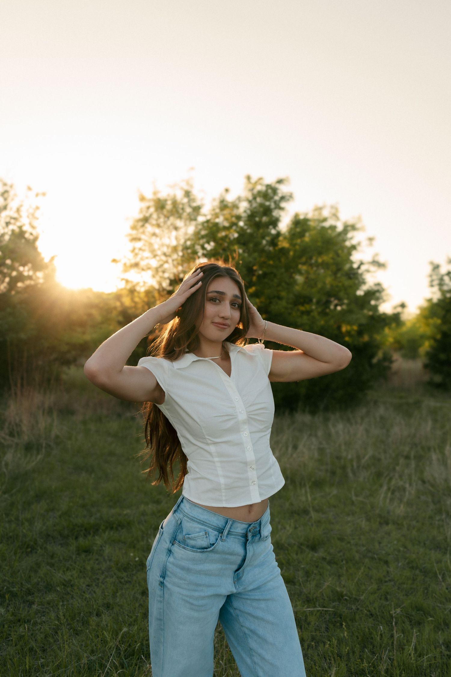 Young woman in white sleeveless top and jeans fixing her hair in a sunlit green field at sunset.
